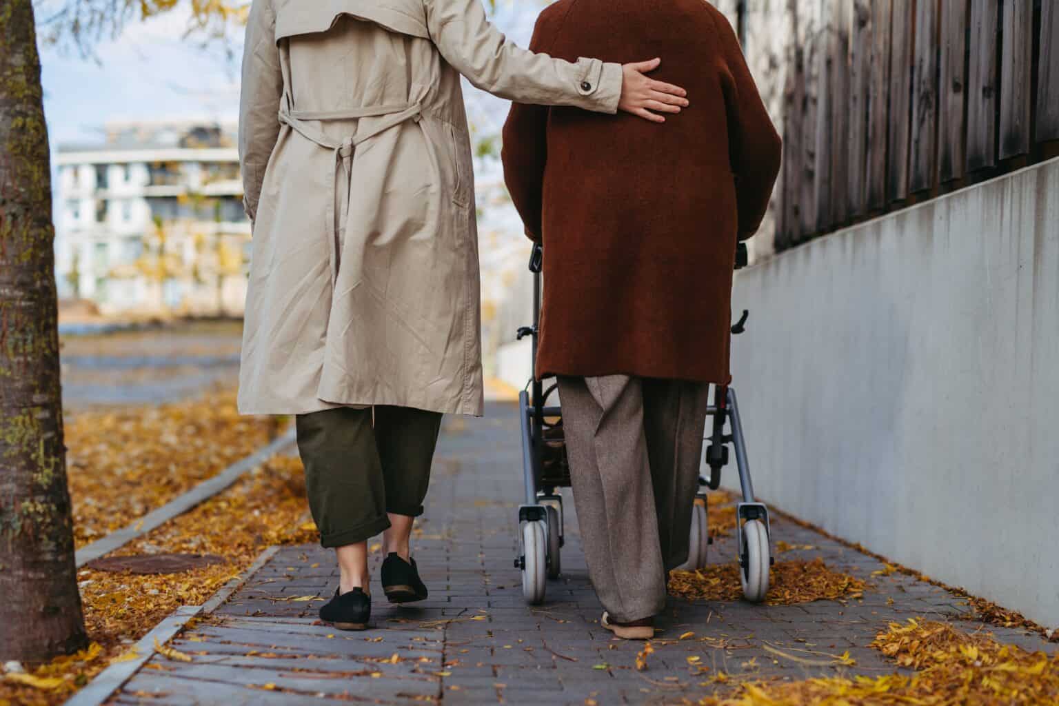 An older person with a walker and a companion walking together along a tree-lined street in autumn